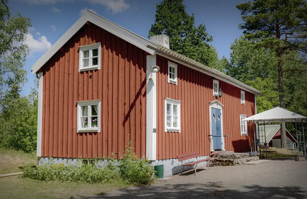 Faluröd stuga i grönskande omgivning.