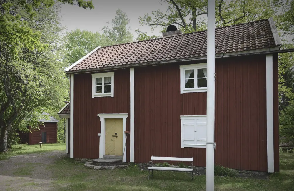 Faluröd stuga i grönskande omgivning.