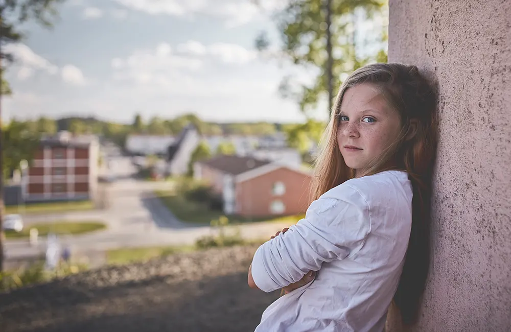 En ung tjej som står lutad mot en vägg i betong. Utsikt över ett bostadsområde finns i bakgrunden.