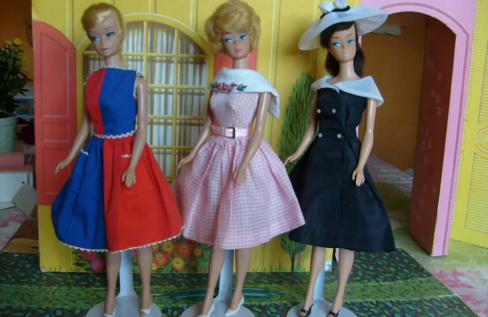 Närbild på 3 barbiedockor uppställda på en stödställning, klädda i olika klänningsstilar och uppställda framför en gul barbiehus vägg