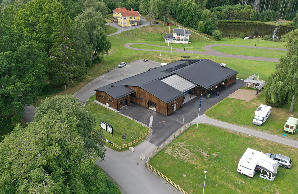 Drönarbild över byggnad med svart tag och träpanel. Till vänster syns tröd och byggnades är omringad av en gräsmatta. Till höger står ett par husbilar parkerade. I bakgrunden syns ett gult hus.