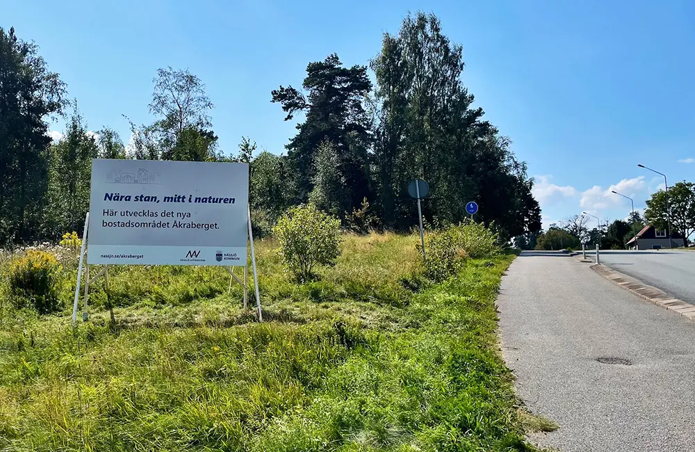 Skylt vid gräs och träd bredvid en gång- och cykelväg med texten "nära stan, mitt i naturen" informerar om att bostadsområdet Åkraberget växer fram.