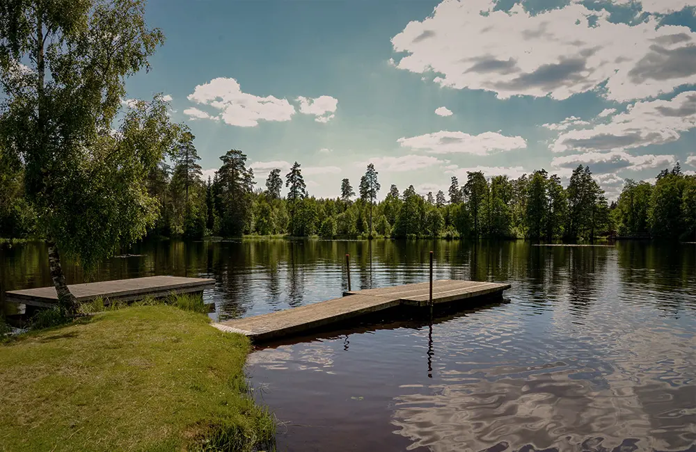 En mindre strand vid sjö. Sjön är spegelblank och från stranden går en brygga ut i sjön.