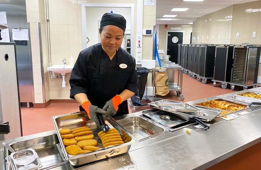 En kvinna arbetar i ett skolkök. Hon har svarta kläder, huvudbonad som skyddar håret och handskar. Hon serverar schnitzel från ett bleck