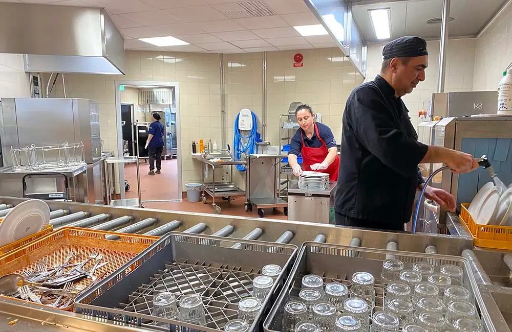 En kvinna och en man arbetar i diskrummet i en skolmatsal. Han sköljer tallrikar och hon kör tillbaka ren disk till rätt station.