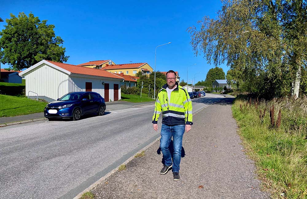 En man i jeans och gul
varseljacka promenerar på trottoaren intill en gata. Det är soligt och blå
himmel. I bakgrunden syns gula bostadshus med rött tak och parkerade bilar i
olika färger.