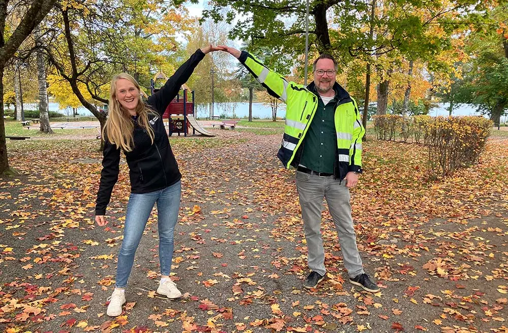 En man och en kvinna står i en park med träd och lekredskap. Det är höst och löven är gula. De ler och tittar in i kameran. Hon har långt hår och svart jacka. han har skägg och gul varseljacka. Med armarna formar de en båge som ska symbolisera en ny entré in i parken.