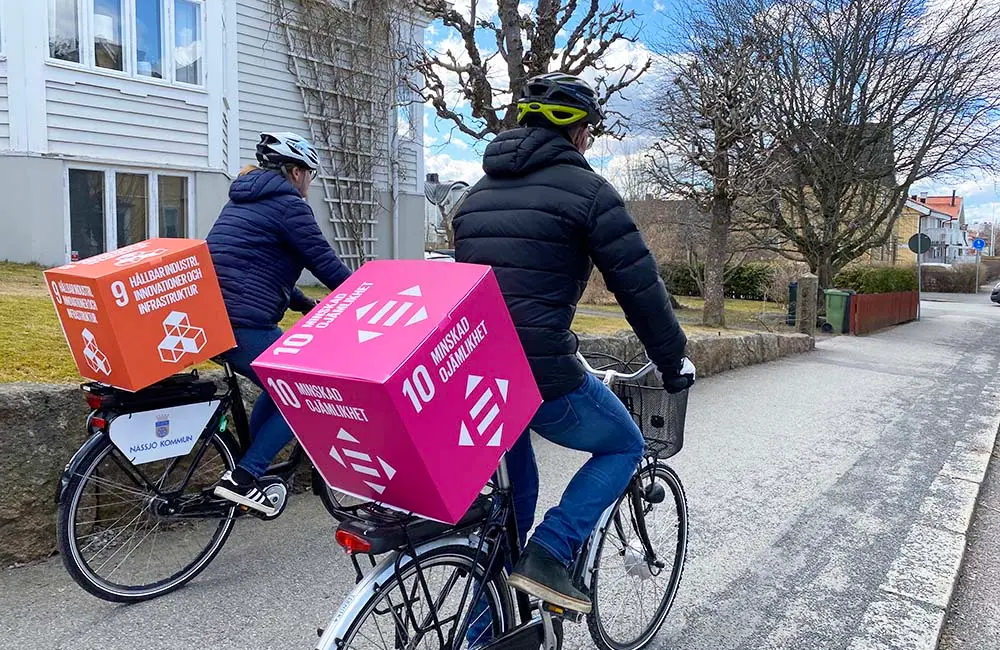 Två personer cyklar med kuber på pakethållaren. Kuberna symboliserar de globala målen 9 (Hållbar infrastruktur) och 10 (Minskad ojämlikhet).