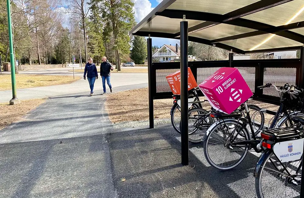 Två personer promenerar på väg mot ett cykelställ med tak. I kuren står två cyklar med globala målen-kuber på pakethållaren. På en av cyklarna syns logotypen Nässjö kommun.