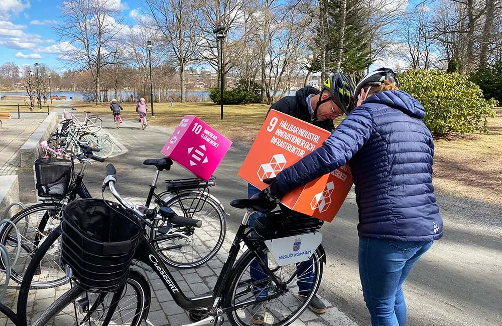 Två personer monterar på globala målen-kuber på pakethållaren till två cyklar. De har hjälmar och det syns en parkmiljö i bakgrunden.