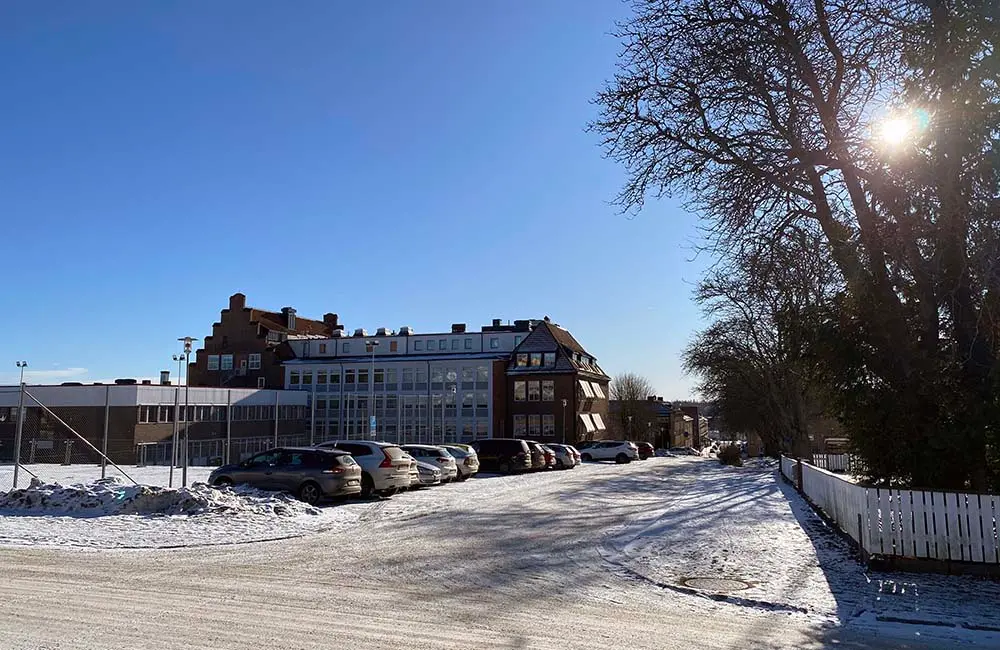 Bilden föreställer en gata som
passerar en skolbyggnad. På gatan står många bilar parkerade. Det är vinter och
vackert väder. Solen skiner och himlen är blå.