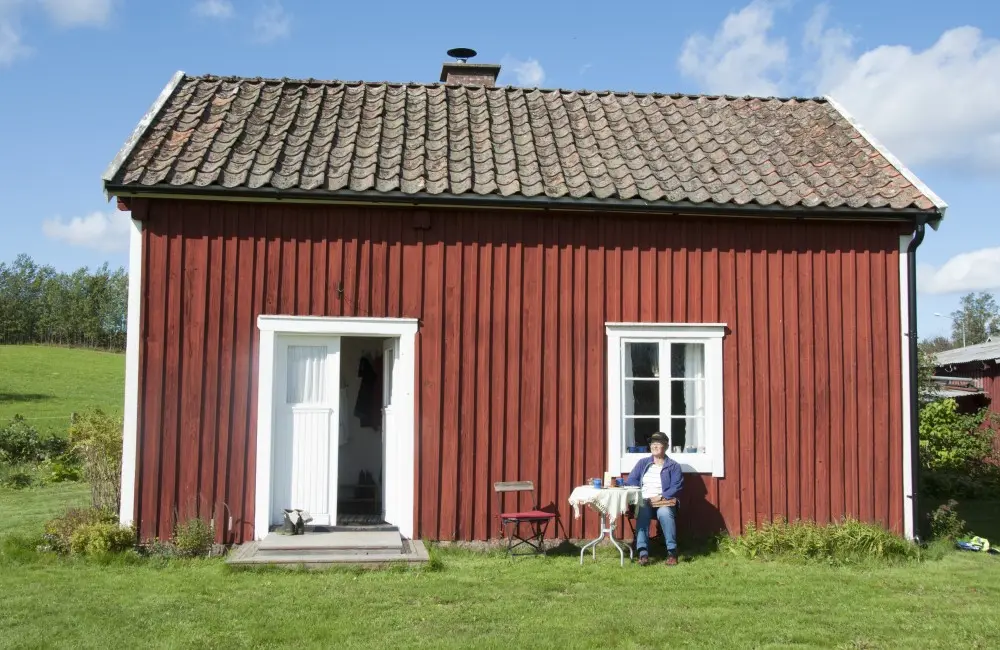 Sommarbild på röd enkelstuga med vita knutar och tegeltak. 