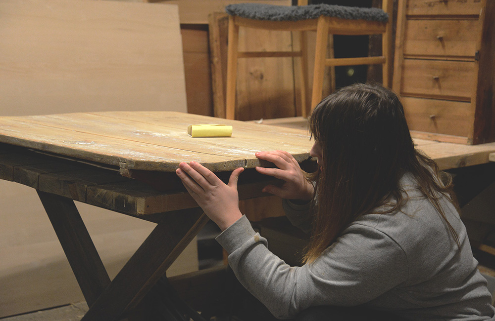 Magdalena sitter på huk och slipar på ett gammalt bord i verkstaden.