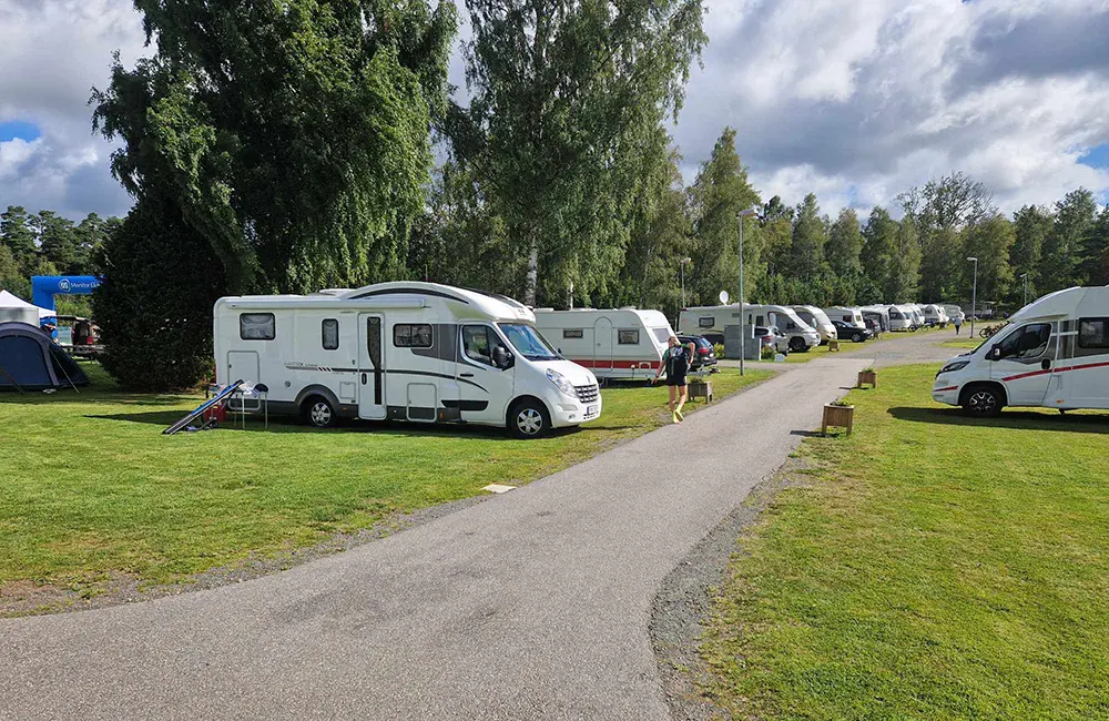 Camping med asfalterad väg i mitten av de vita husbilarna och husvagnarna på rad. Gräset är grönt på sidorna och det är soligt väder. I bakgrunden syns stora träd.