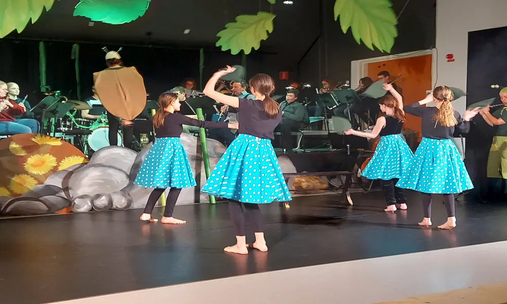 På scenen spelar ungdomsblåset. Dirigent är "fästingen". På golvet framför scenen dansar 4 tjejer i turkosa kjolar med vita prickar.