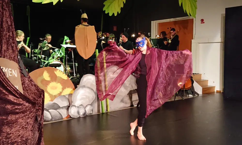 I bakgrunden på scenen
sitter ungdomsblåset och spelar. Framför scenen är en dansare utklädd till en
vacker fjäril med violetta vingar och en blåglittrig mask för ögonen.