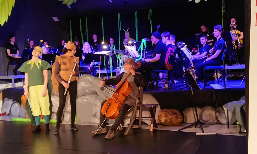 I bakgrunden syns orkestern i ett blått strålkastarljus. Framför scenen står "syrsan" och "fästingen" och pratar med spindeln som spelar på sin cello.