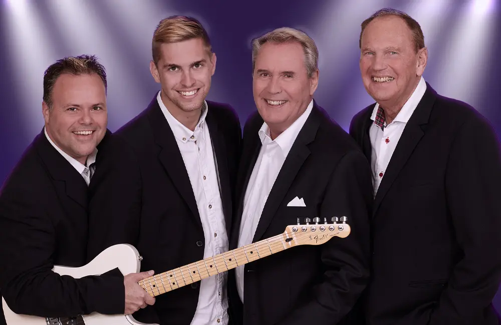 Fyra män i svarta kavajer . En av männen håller i en basgitarr.