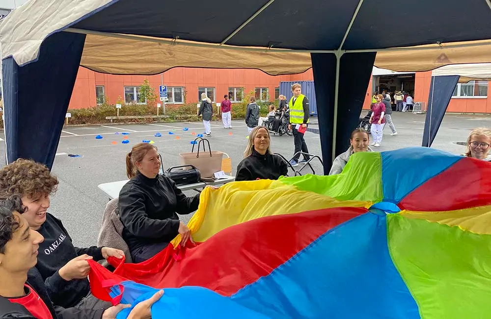 En grupp människor sitter i ring i en mässmonter och övar samarbete med ett stort regnbågsfärgat tygstycke.