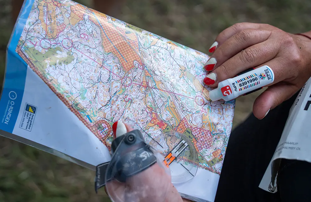 Två händer med rött och vitt nagellack håller i en orienteringskarta. Personer håller även i ett förstoringsglas och en markör finns på ena pekfingret. 