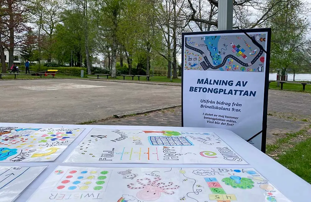 Teckningar ligger på ett bord framför en asfalterad yta. I bakgrunden syns en skylt med ett färglatt och fantasifullt motiv som informerar om att betongplattan ska målas.