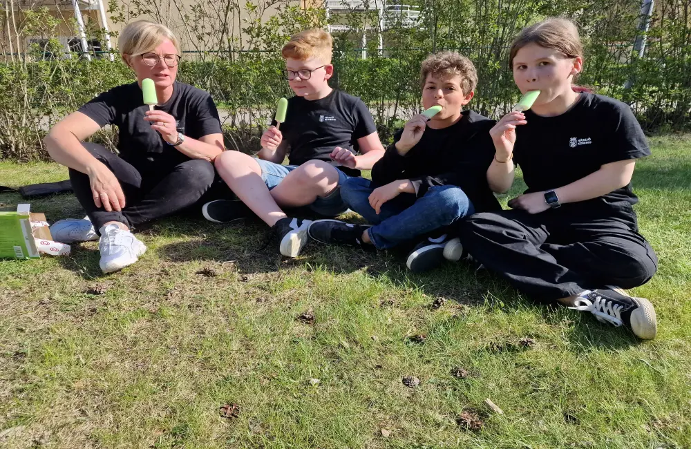 En kvinna och tre barn sitter på en gräsmatta och äter en grön isglass. De är klädda i likadana svarta T-shirts och har lång- respektive kortbyxor på sig. I bakgrunden syns en grönskande häck.