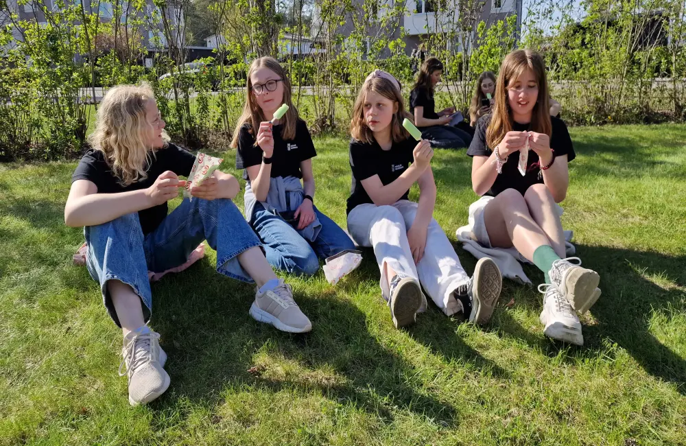 Fyra tjejer sitter på en gräsmatta och äter en grön isglass. De är iklädda likadana svarta T-shirts och har lång- respektive kortbyxor på sig.