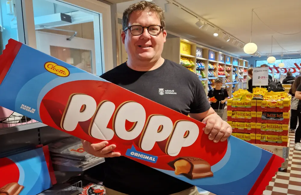 En leende man med kort rödbrunt hår och glasögon, står i en butik och håller upp en enormt stor kartong som det står Plopp på. I bakgrunden skymtas mer godis.