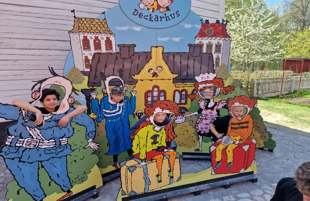 Vid en färgglad tavla föreställande en massa hus och människor , sticker några barn ut sina huvuden i små hål i tavlan. Längst upp på tavlan står det deckarhus.