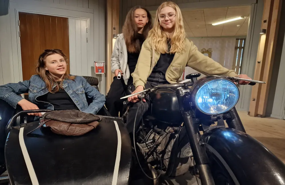 På en motorcykel med sidovagn sitter tre tjejer. Två sitter på motorcykeln och en sitter i sidovagnen.