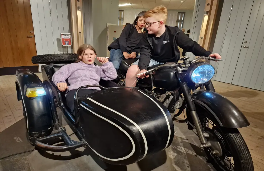 På en motorcykel med sidovagn sitter tre barn. Två killar sitter på motorcykeln och en tjej sitter i sidovagnen.
