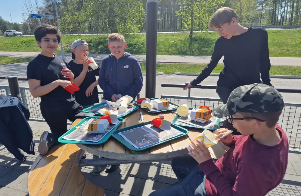 Fem killar sitter och står runt ett picknickbord. På bordet syns brickor med hamburgare och läskmuggar.