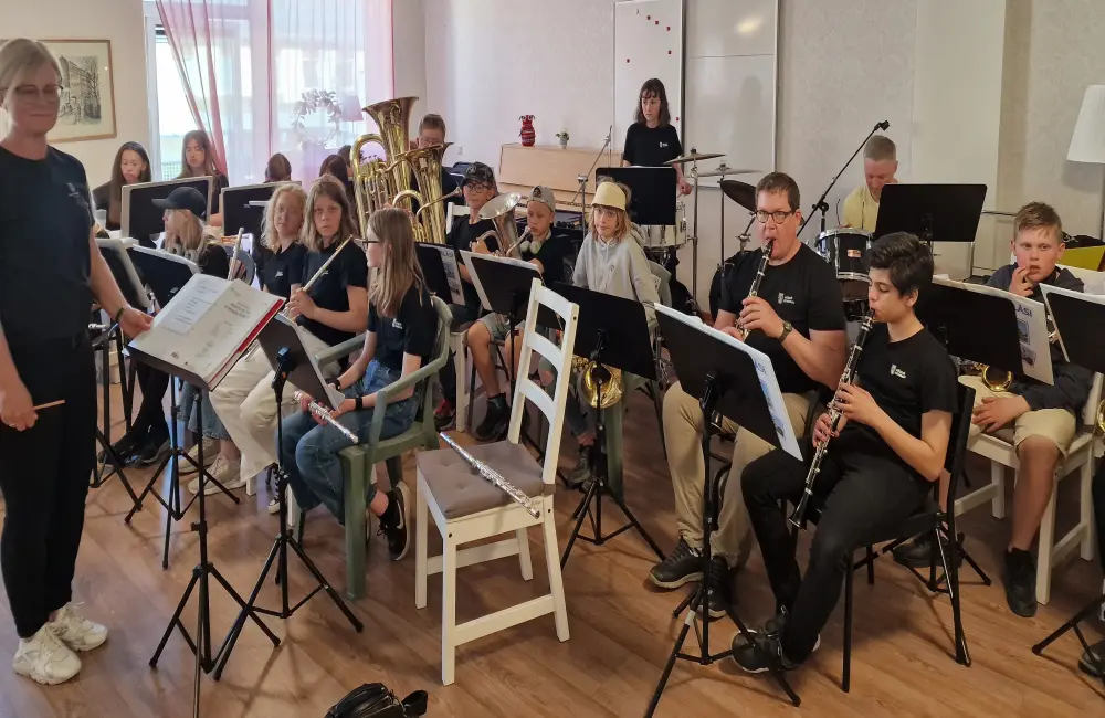 Man ser en blåsorkester som består av barn i mellanstadieåldern. De sitter på stolar med sina instrument. Framför dem står en kvinna vid ett notställ. Man kan även skymta några vuxna i orkestern.