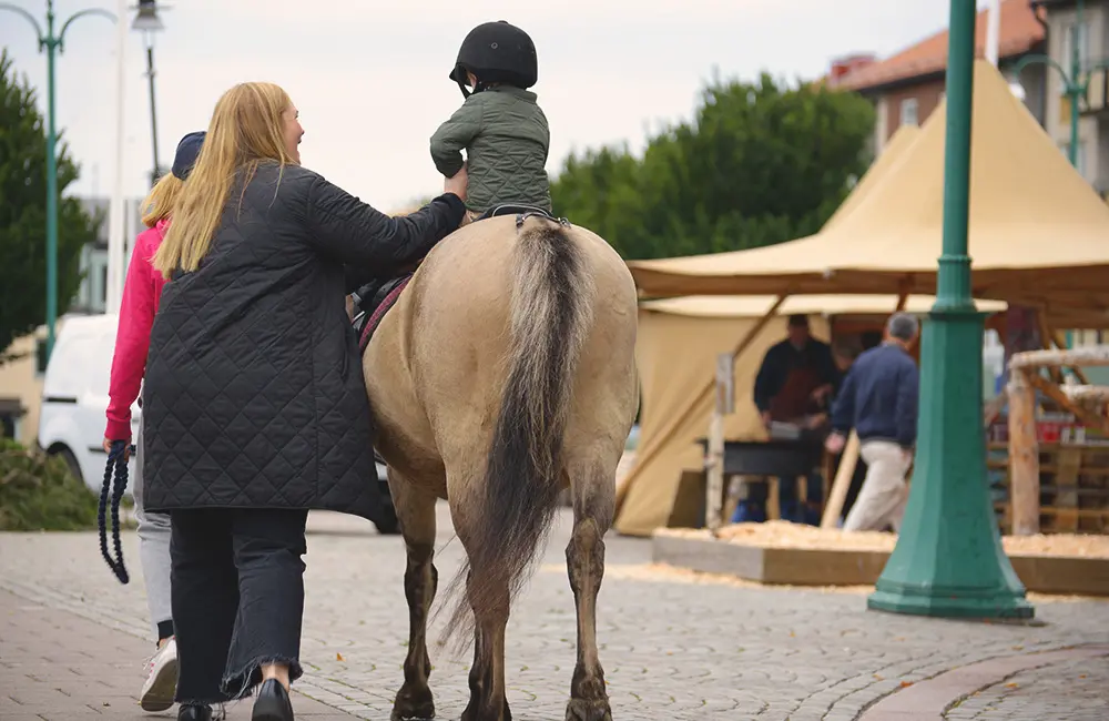 Litet barn sitter på en stor häst. Barnets mamma går bredvid och håller i.