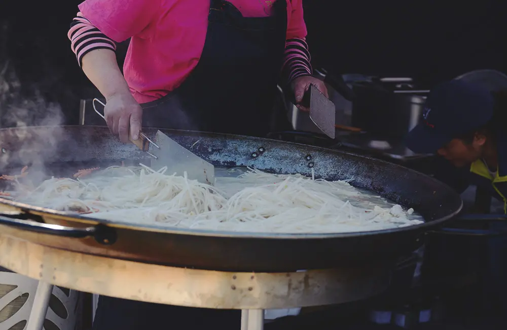 En stor muurikka med nudlar och wok. Det ryker om pannan och en person i rosa kläder står och rör runt i maten.