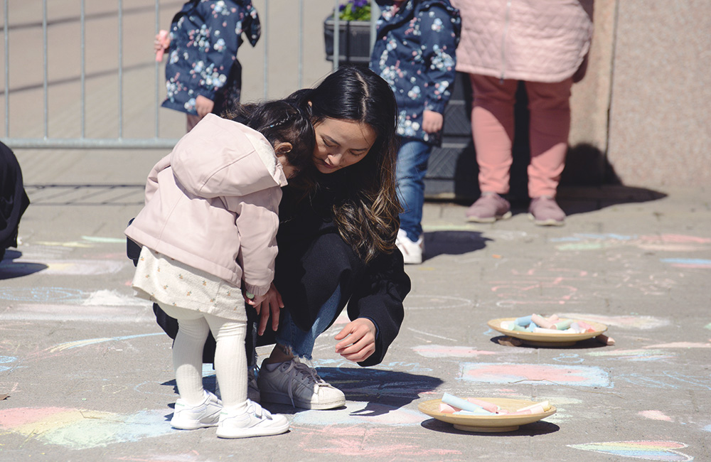 Ett barn och en vuxen står vid en skål med asfaltskritor och ritar på asfalten intill med kritorna.