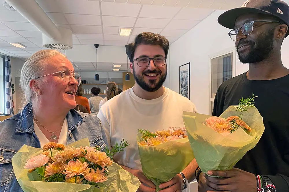 Tre personer, en äldre kvinna och två unga män poserar för kameran med stora blombuketter. De ser glada ut.