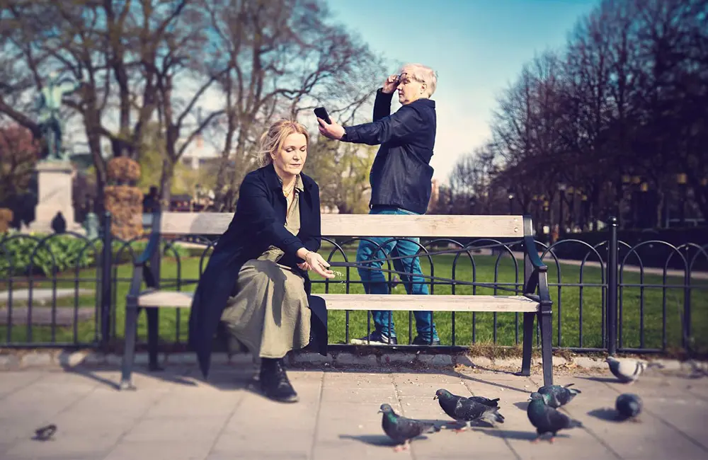 En kvinna sitter på en parkbänk. Hon matar duvor. Bakom parkbänken står en man i glasögon och tittar i sin mobil.