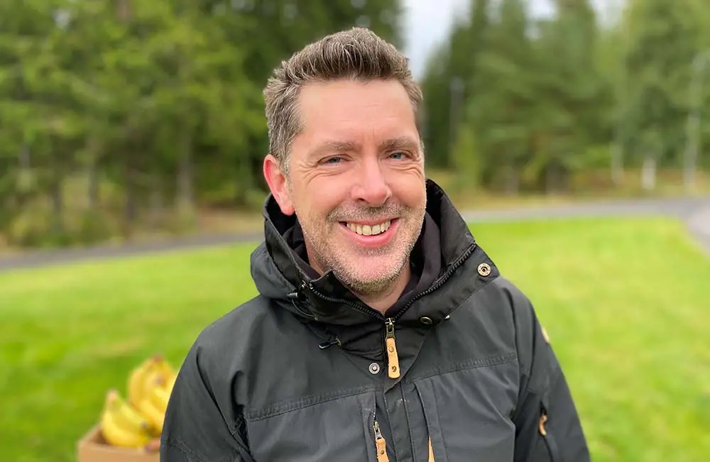 En man i höstjacka ler in i kameran. I bakgrunden syns en låda bananer.