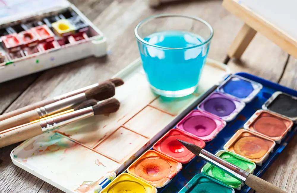 Vattenfärgspalett med olika penslar och ett vattenglas med blå färgat vatten i. Alla saker ligger på ett träbord. 