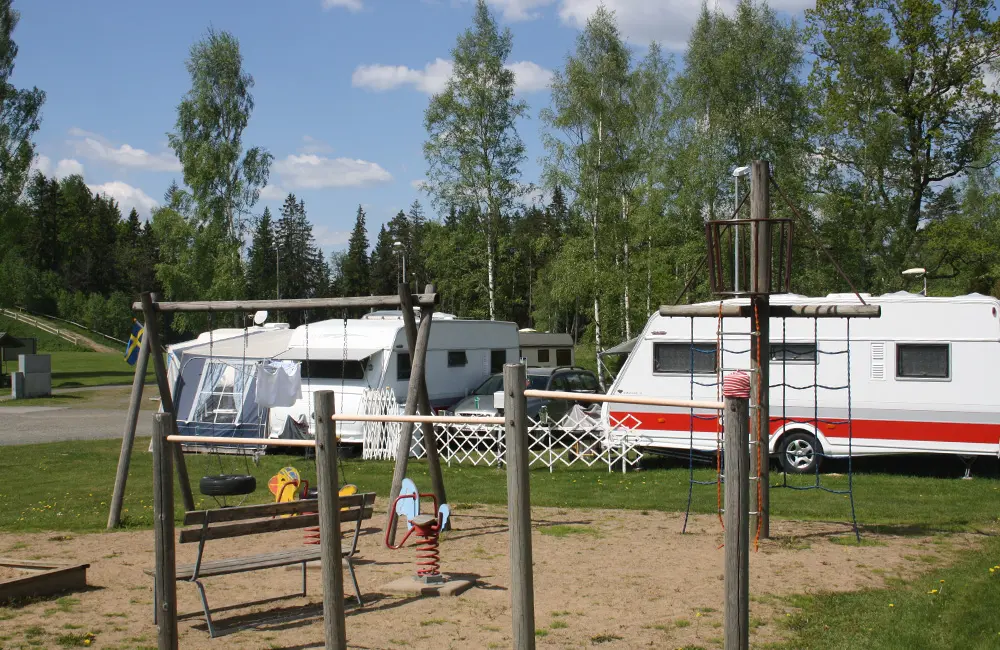 En bild på en lekplats med olika lekställningar. I bakgrunden syns ett par husvagnar. 