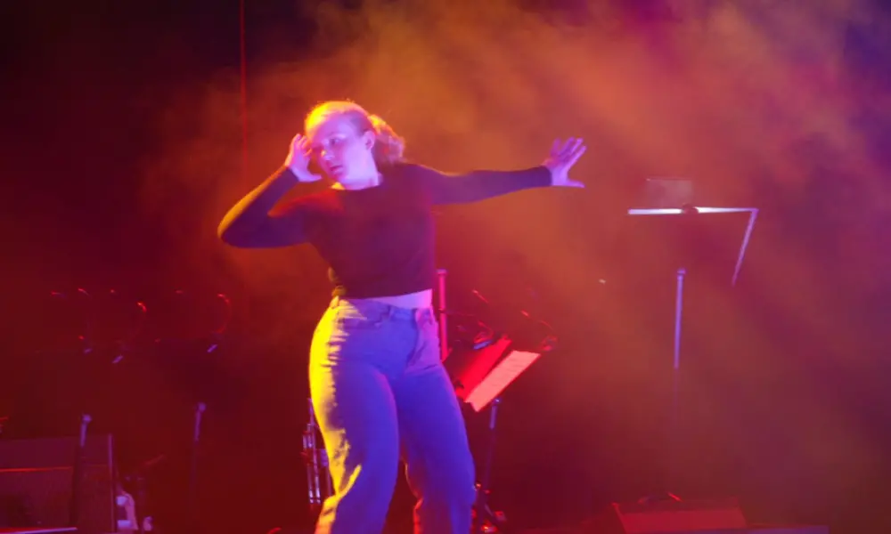 I ett rött sken dansar en tjej på scenen. Hon ser sig åt sidan och håller den ena armen böjd och den andra är utsträckt åt motsatt sida. Hon är klädd i svart tröja och blå jeans. Håret är uppsatt i en tofs. Bakom henne syns ett notställ.