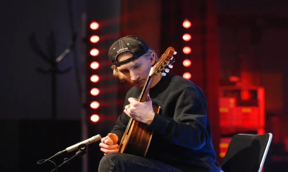 På en stol sitter en ung kille med en akustisk gitarr i knät. Framför gitarren finns en liten mikrofon. Killens klädsel är svart och han bär en bakåtvänd keps. I bakgrunden syns ett par stativ med lampor.