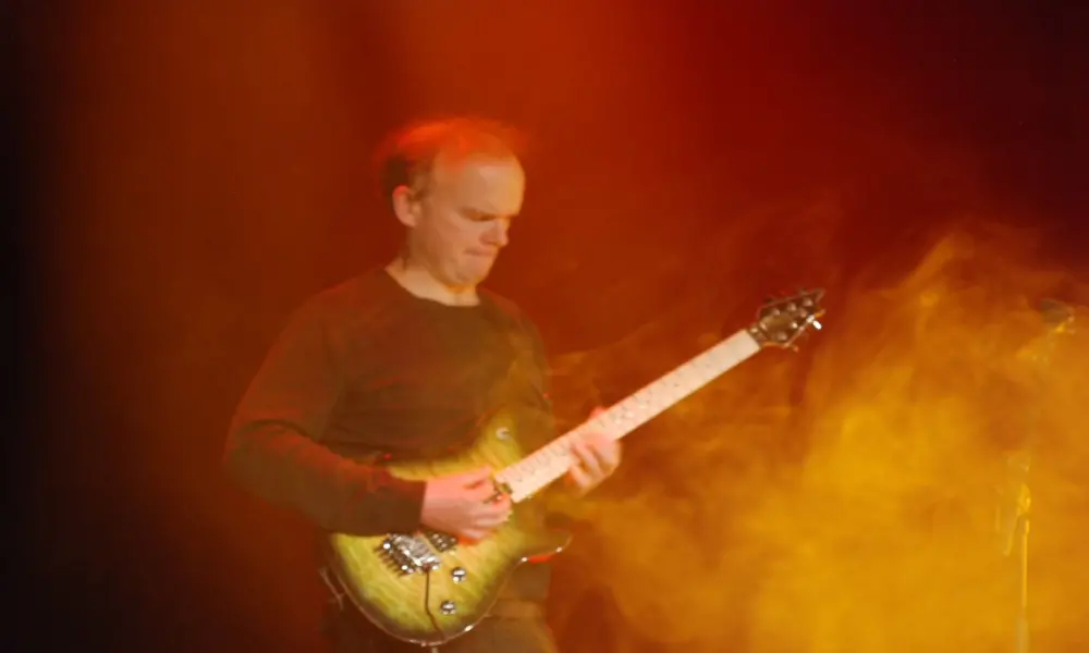 I ett moln av rök och röd-gult sken står en man och spelar på en gul elgitarr. Han bär svarta kläder.