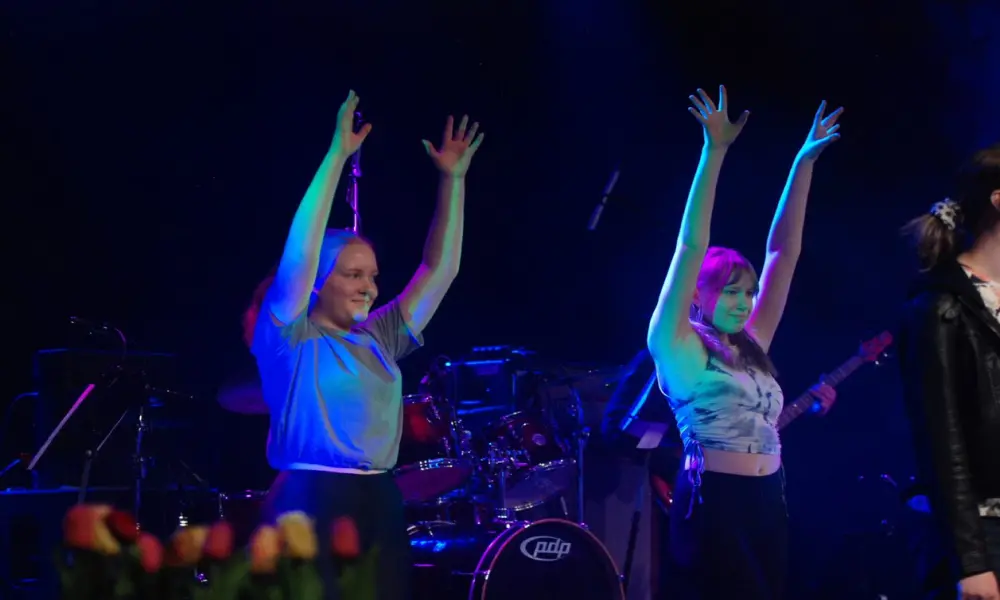I ett blålila sken står två glada tjejer och dansar med sina armar uppsträckta. De har grå tröjor på sig och svarta byxor.