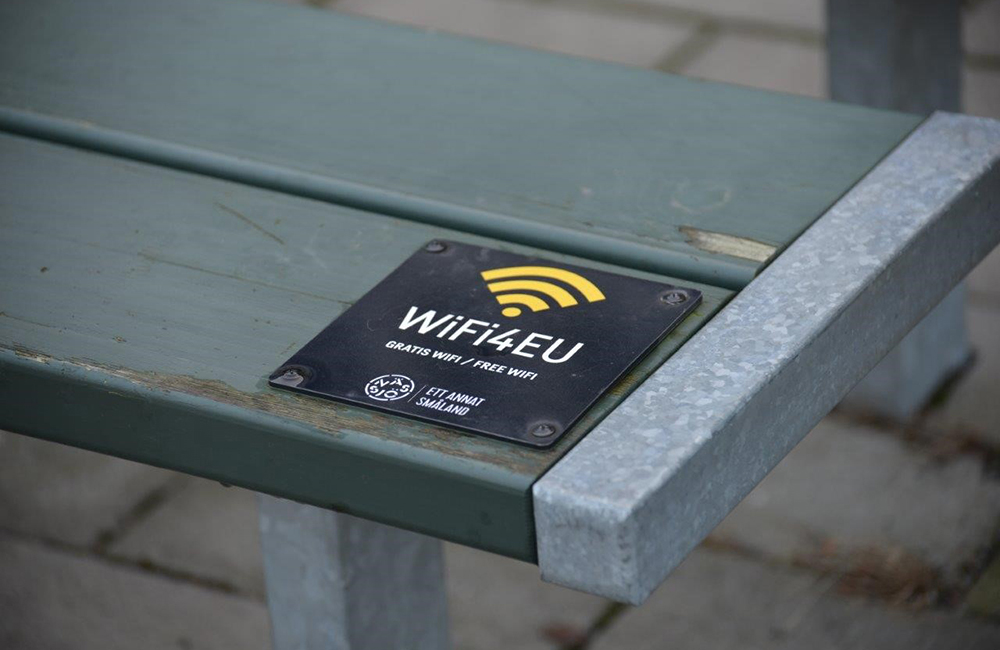 Ytterkanten på en grågrön parkbänkssits där en svart liten fyrkantig skylt med vita bokstäver visar WiFi4EU som är Nässjö shoppings erbjudande om fritt wi-fi till besökare i centrala delar av kommunorterna.