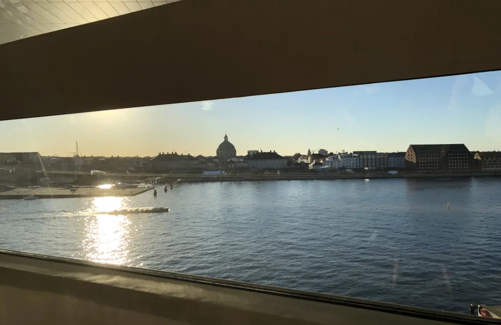 Bilden är tagen genom ett fönster . Utanför finns vatten och i bakgrunden tornar hus upp sig. Solen skiner.