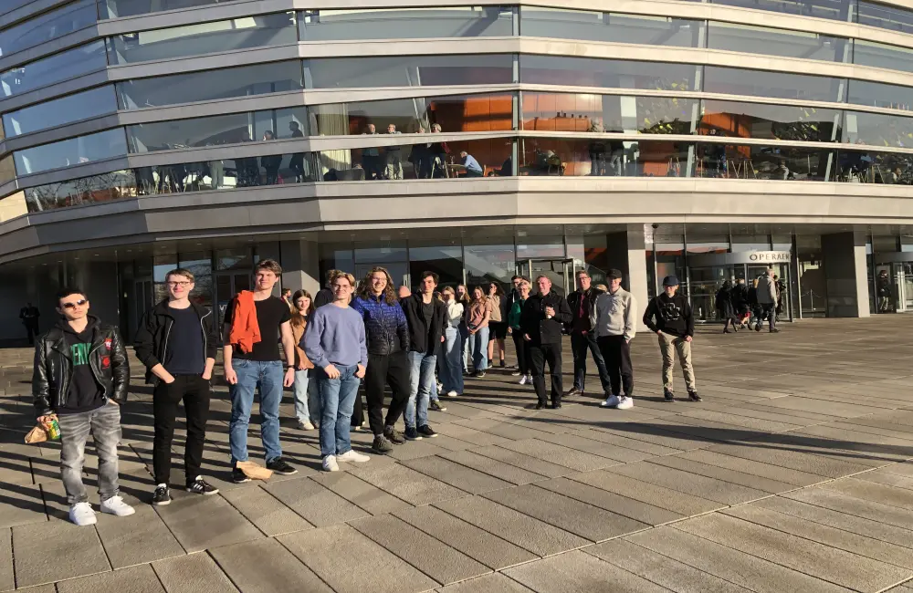 På bilden står en grupp ungdomar samlade utanför en stor byggnad, där fasaden är gjord av fönster.