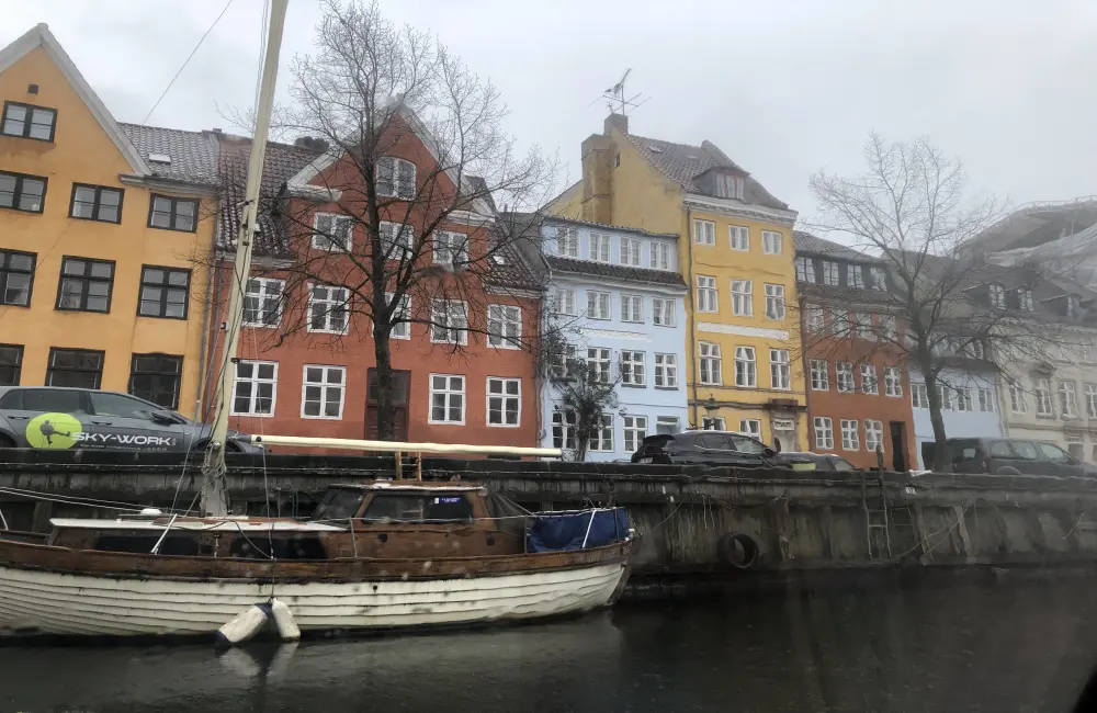 Bilden visar en större träbåt som ligger förtöjd. Bakom syns flervåningshus i olika färger.