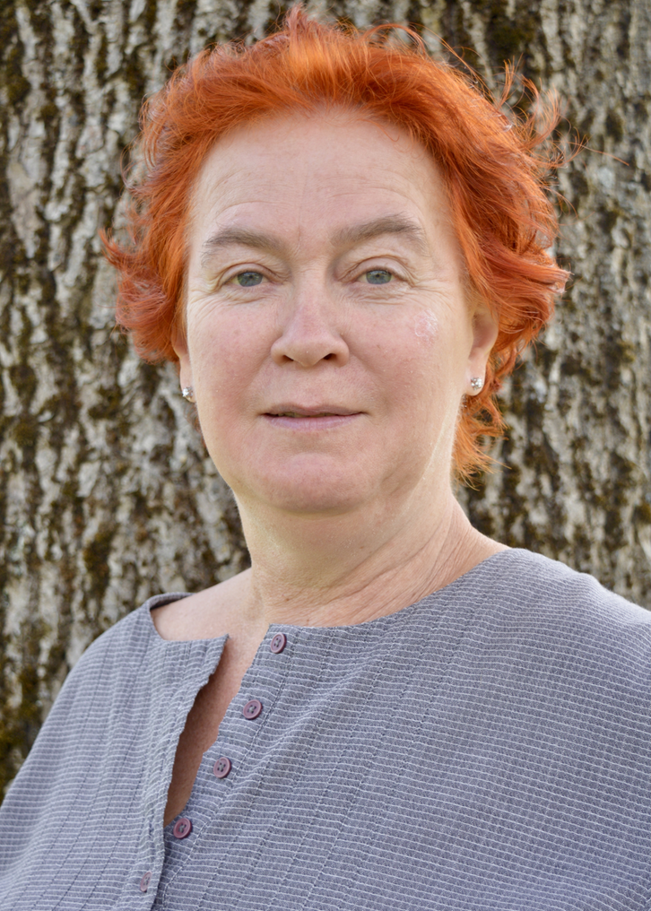 En kvinna med kortklippt rött hår. På sig har hon en blågrå linneblus med knappar. Bilden är tagen utomhus framför en trädstam.